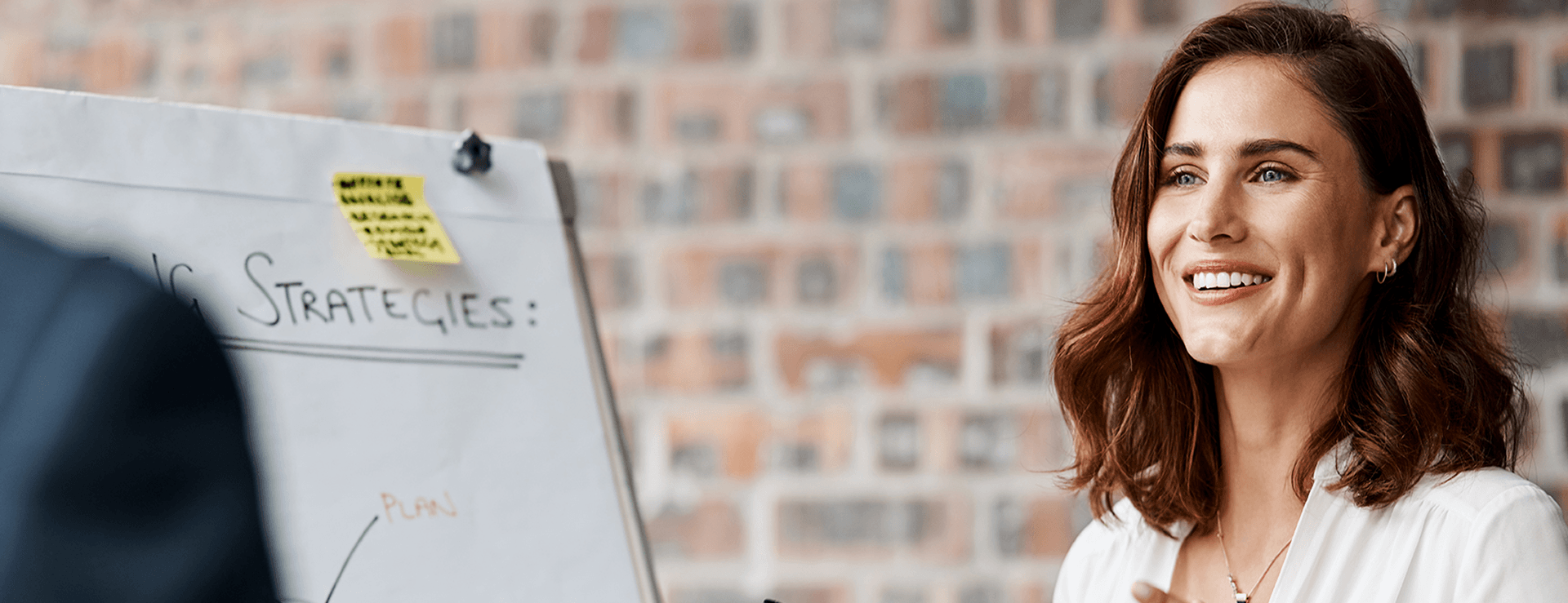 Close-up of a smiling brunette businesswoman in a white shirt talking during a presentation next to a flipchart titled "Strategies" in front of a brick wall.