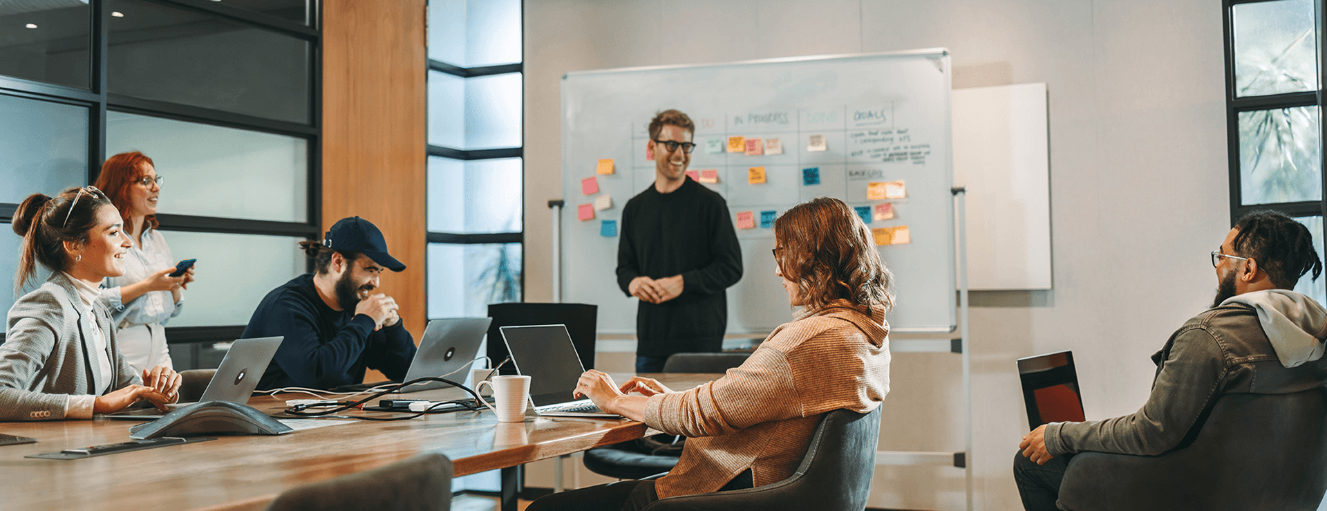 A diverse group of colleagues hold a casual meeting in a modern meeting room; one man smiles while standing in front of a whiteboard with colorful sticky notes, and the others sit around a table with laptops.
