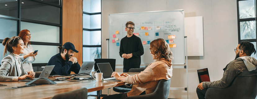 A diverse group of colleagues hold a casual meeting in a modern meeting room; one man smiles while standing in front of a whiteboard with colorful sticky notes, and the others sit around a table with laptops.