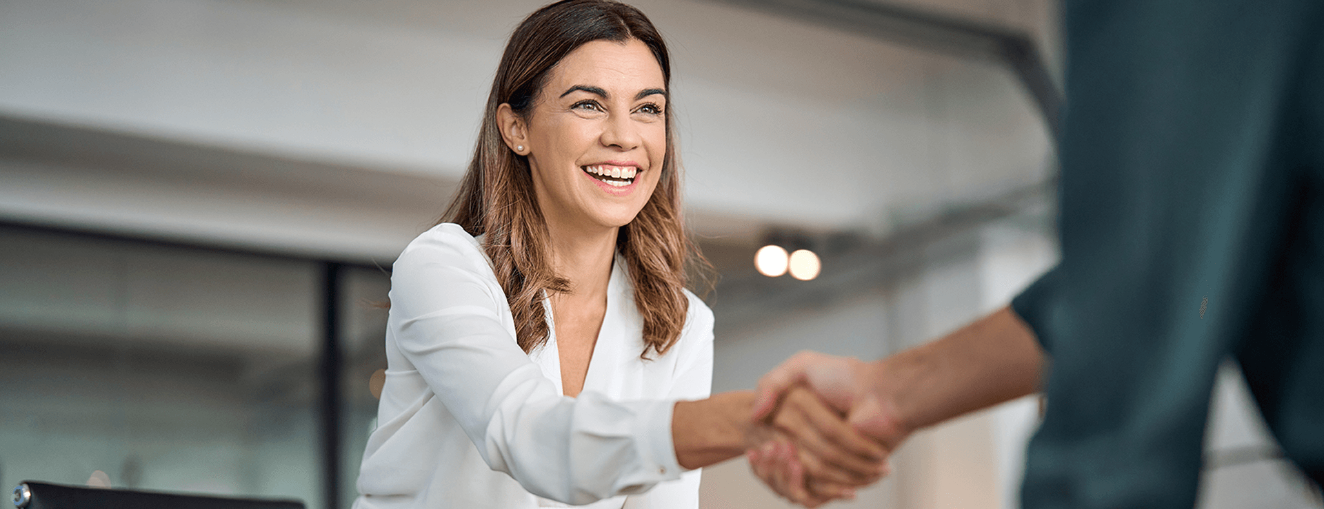 A smiling businesswoman in a white blouse shakes hands with a business partner in a bright office.