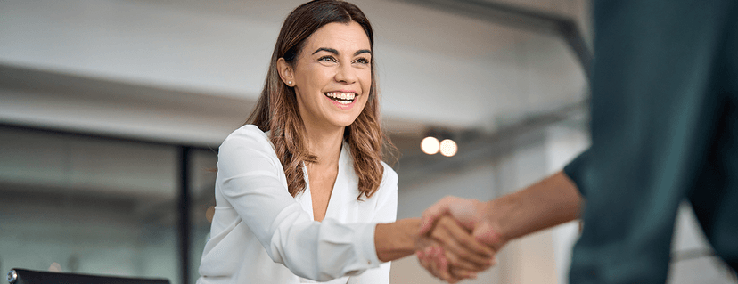 A smiling businesswoman in a white blouse shakes hands with a business partner in a bright office.