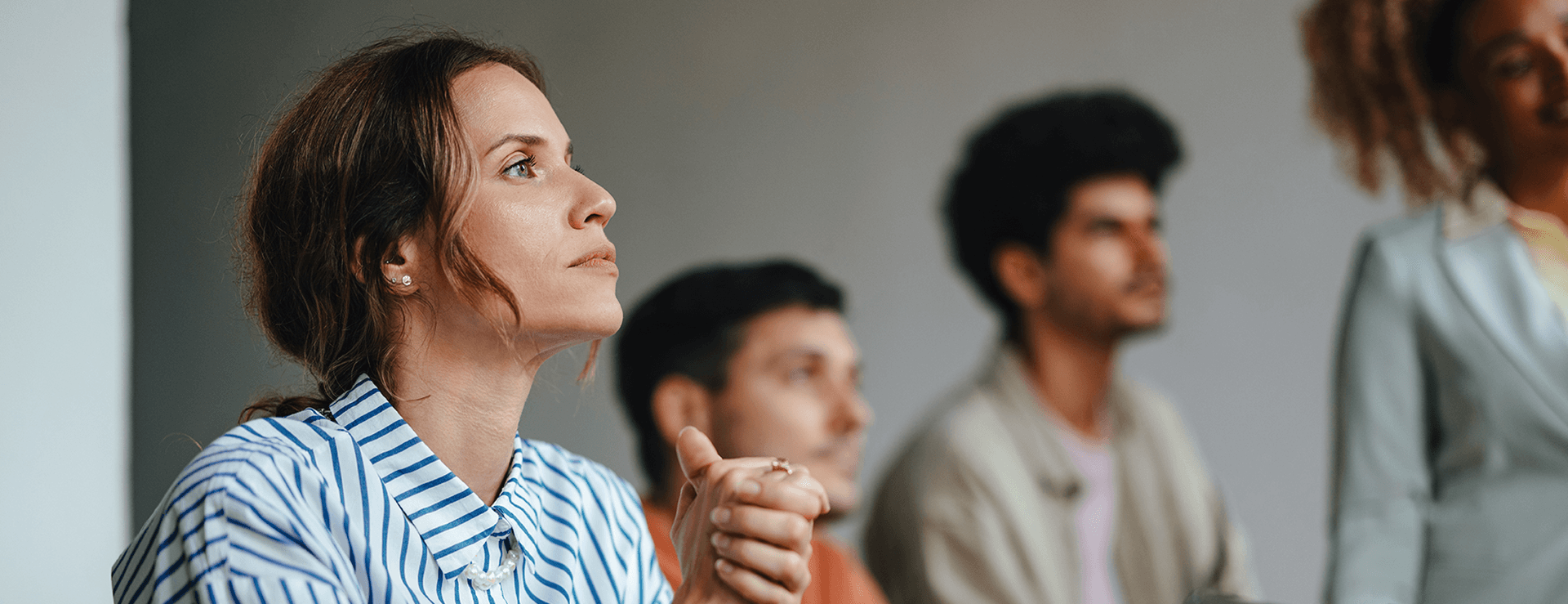 Focus on an attentive woman in a blue and white striped shirt sitting in an audience, hands folded and looking intently at the speaker (partially visible on the right). Other spectators are blurred in the background.