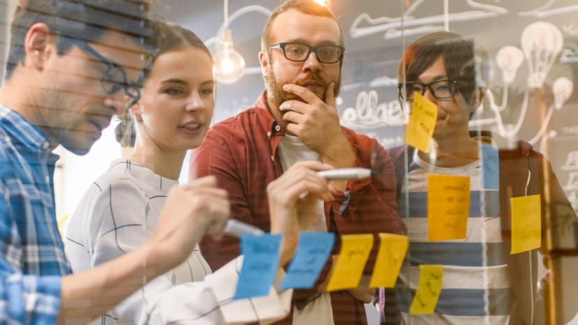 Young woman in a gray sweater presents to a group of colleagues behind a glass wall and points to a whiteboard with post-its.