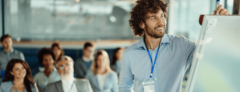 Smiling trainer with curly hair and blue shirt writes on a whiteboard in front of a group of attentive participants.