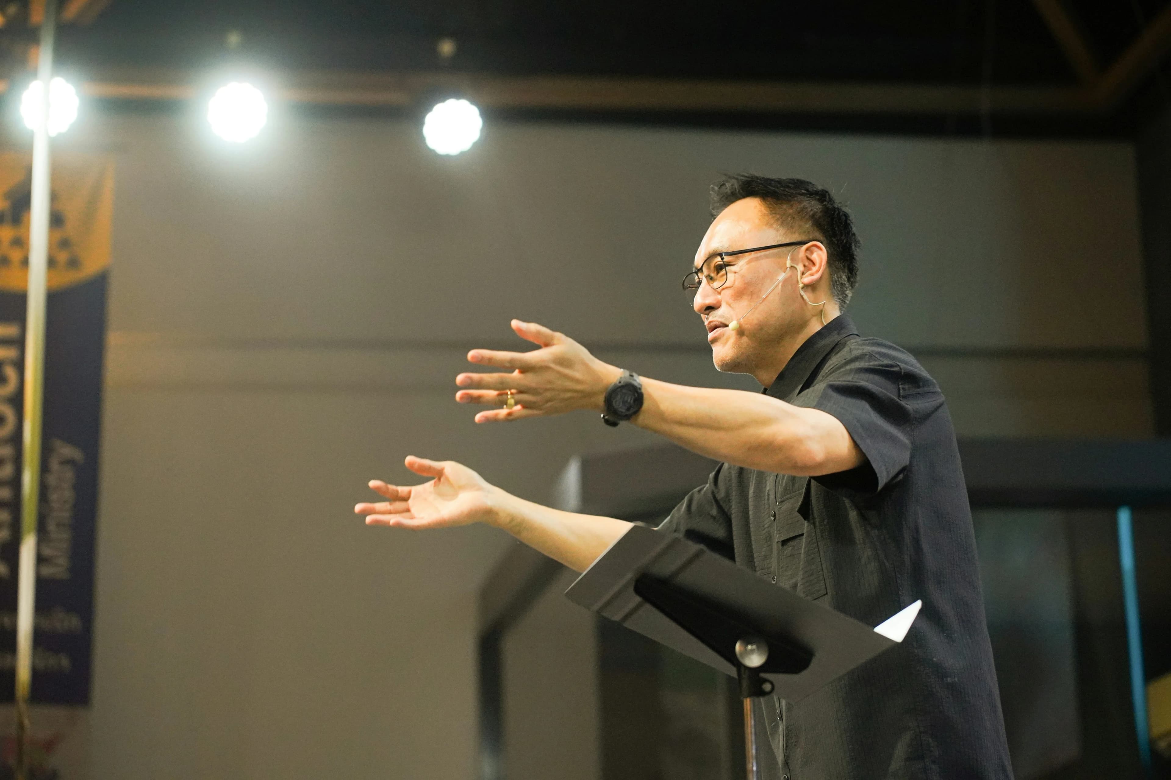 An adult man wearing glasses and a black button-down shirt speaks passionately with open arms at a podium under bright stage lights.