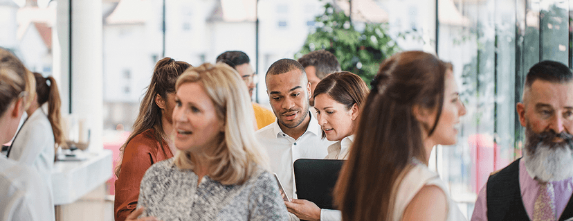 Close-up of a busy group of people networking and chatting at an event; a man and a woman in the center are looking at a laptop together.
