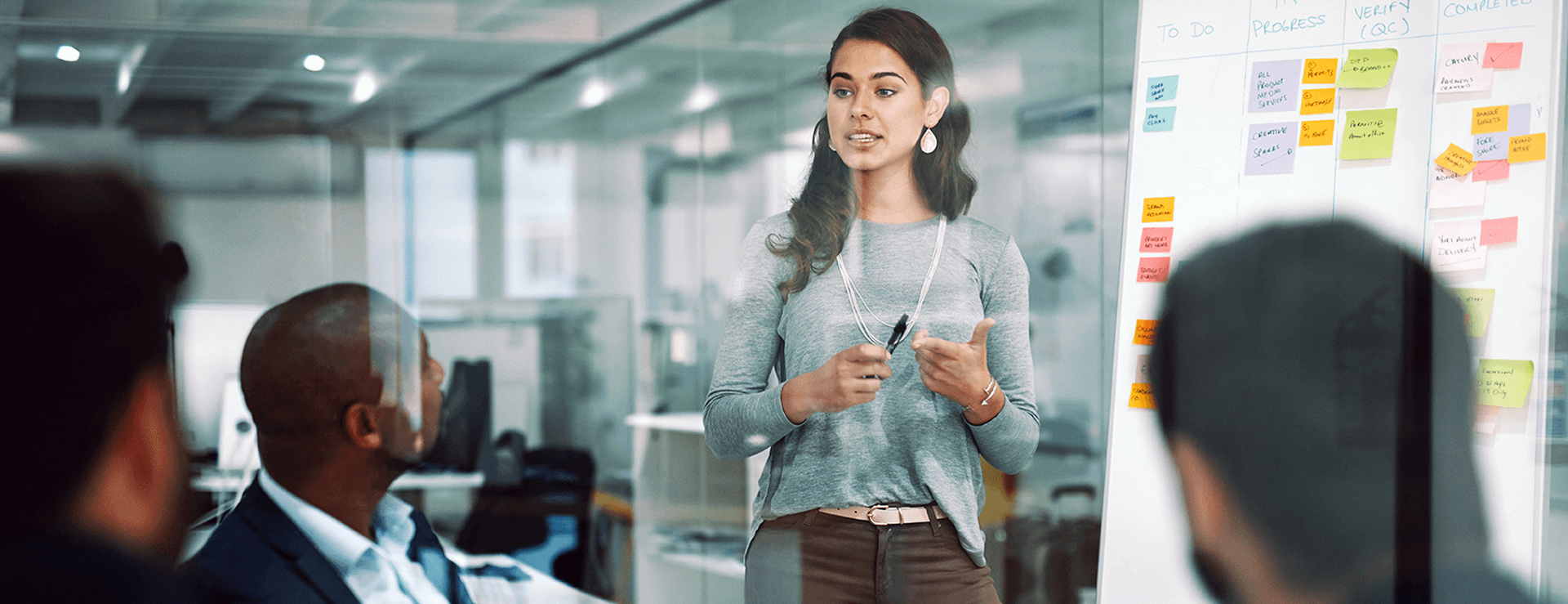 Dark-haired woman in a gray sweater holds a pen and speaks in front of a group of participants while standing at a whiteboard with post-its.