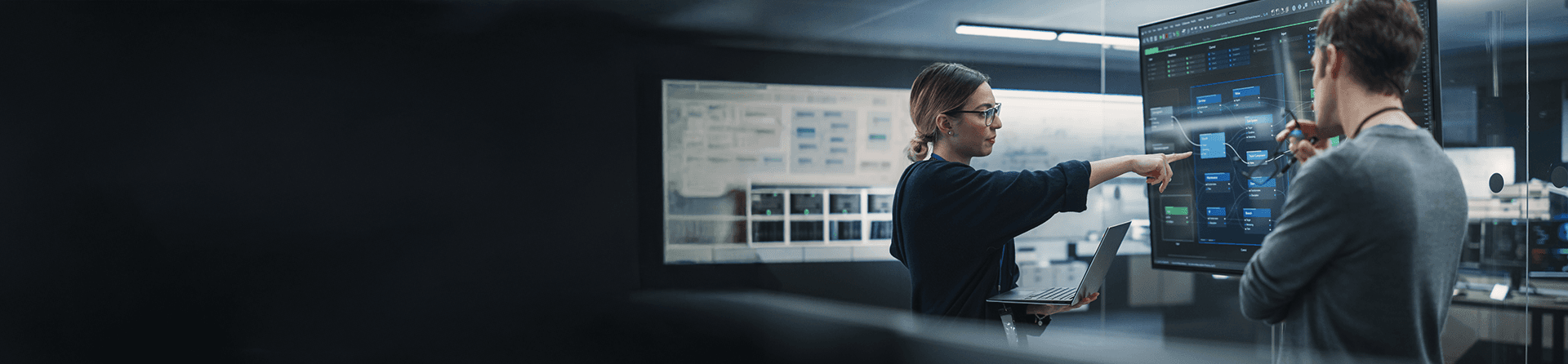 Woman with glasses and laptop points to a large data chart.
