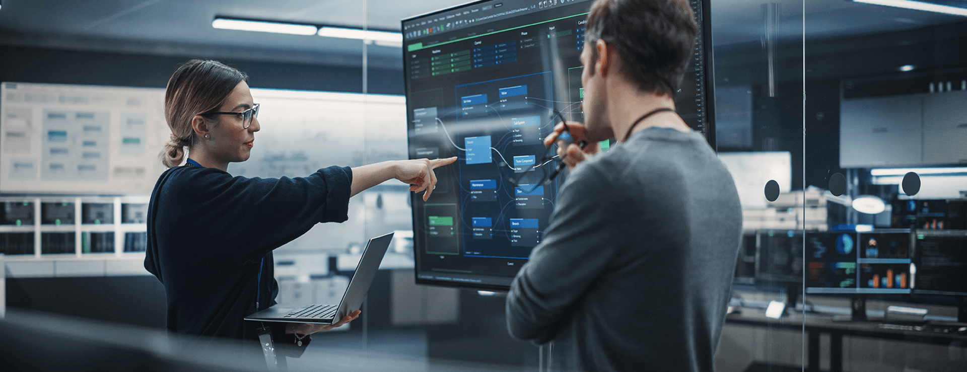 Woman with glasses and laptop points to a large data chart.