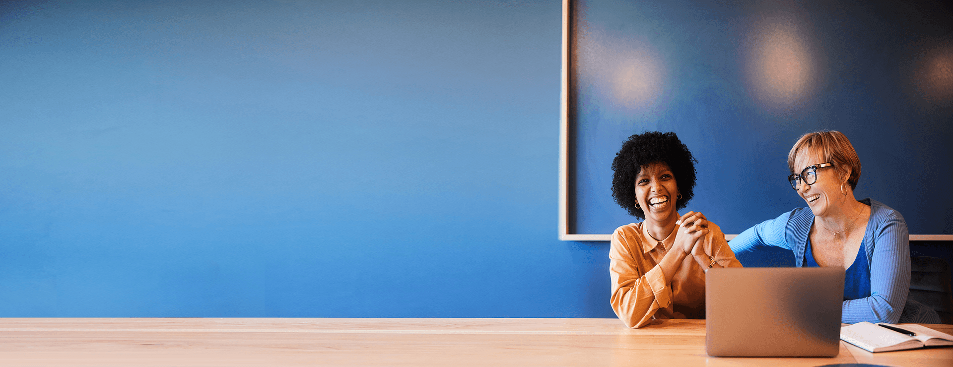 Two smiling women, one young and one older, sit at a wooden table in front of a bright blue wall with a laptop and notebook.