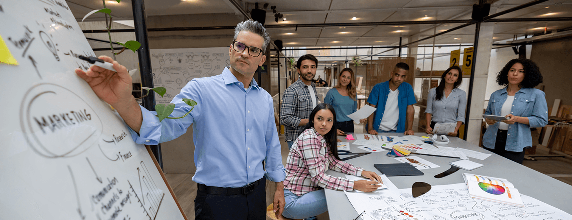 Middle-aged man with glasses in a blue shirt writes MARKETING on a whiteboard while his diverse team looks on in the modern office.