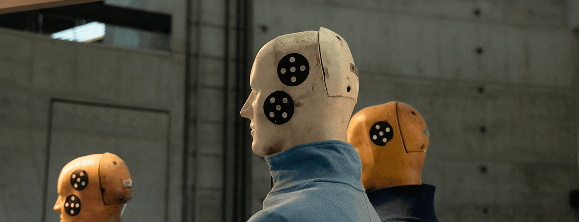 Nahaufnahme der Köpfe von drei Crashtest-Dummys in einem Labor; der weiße Dummy in der Mitte trägt einen blauen Kragen und hat schwarze, kreisförmige Messpunkte im Gesicht.